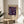 May include: Framed poster featuring Yayoi Kusama's colorful floral infinity artwork with purple background and multicolored daisy-like flowers, displayed on a light wood round side table next to a beige upholstered chair. Natural sunlight streams across the neutral-toned room with white walls and a plant visible in the corner.