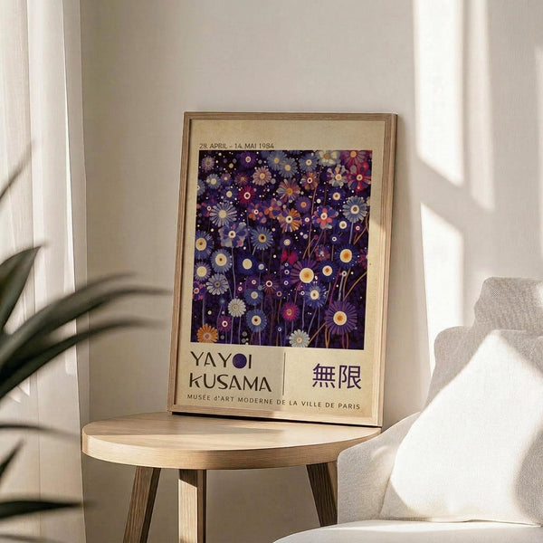 May include: Framed poster featuring Yayoi Kusama's colorful floral infinity artwork with purple background and multicolored daisy-like flowers, displayed on a light wood round side table next to a beige upholstered chair. Natural sunlight streams across the neutral-toned room with white walls and a plant visible in the corner.