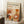 May include: Framed art print leaning against white wall corner on textured cream rug. Print features abstract botanical design with orange, rust, and cream organic shapes, circles, and stylized plants. Text reads "YAYOI KUSAMA" with Japanese characters and "JAPAN 1987". Natural wood frame, cozy room setting with coffee cup on wooden block and beige fabric visible.