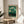 May include: Framed art print with green gradient background featuring whimsical white and golden dotted trees and botanical elements. Text reads "YAYOI KUSAMA" and "KYOTO" in black letters at bottom. Natural wood frame sits on round wooden side table next to beige upholstered chair, with plant leaves visible in corner and soft natural lighting creating shadows on white wall.
