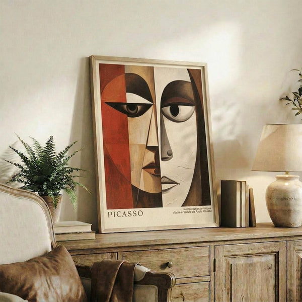 May include: Framed cubist-style portrait print displaying geometric face with orange and brown color blocks, asymmetrical eyes, and angular features in wooden frame. Print sits on rustic wooden sideboard alongside potted fern, ceramic vase, and stacked books in warm-toned living room setting with cream upholstered furniture.