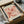 May include: Vintage-style botanical poster with coral and pink flowers in Matisse cut-out style, displayed in black frame inside brown cardboard shipping box with protective tissue paper. Wooden dowels visible alongside for hanging. Cream background with "Berggruen & Cie" gallery text at bottom.