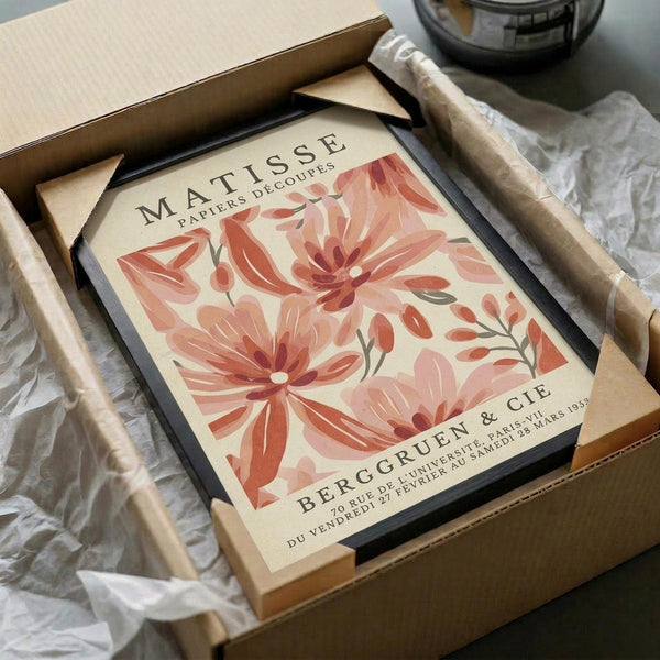 May include: Vintage-style botanical poster with coral and pink flowers in Matisse cut-out style, displayed in black frame inside brown cardboard shipping box with protective tissue paper. Wooden dowels visible alongside for hanging. Cream background with "Berggruen & Cie" gallery text at bottom.