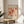 May include: Framed Matisse exhibition poster featuring vibrant orange and pink floral cut-out designs, displayed on a round wooden stool beside a beige upholstered chair. Soft natural lighting creates shadows across white walls, with green plant leaves visible in the corner. The vintage-style poster shows botanical shapes in warm coral tones with French gallery text below the artwork.