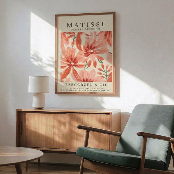 May include: Framed vintage art poster featuring coral and pink abstract floral cutout designs on cream background, displayed above wooden sideboard with table lamp in mid-century modern living room setting with sage green upholstered chair and natural sunlight casting shadows on white wall.