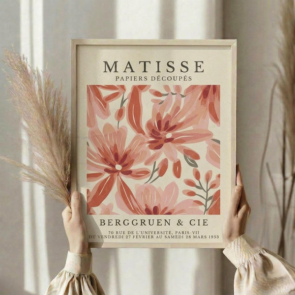 May include: Person holding framed vintage-style art print featuring abstract pink and coral flowers in Matisse's cut-out style, with cream background and exhibition text. Light wood frame, dried pampas grass visible to left, soft natural lighting near white curtained window.