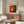 a living room with a large abstract painting hanging on the wall above a wooden cabinet. The painting has a vibrant mix of red, orange, and yellow colors, creating a striking visual contrast. The room also includes a white sofa, a glass coffee table, and a lamp, contributing to the overall modern and stylish aesthetic.
