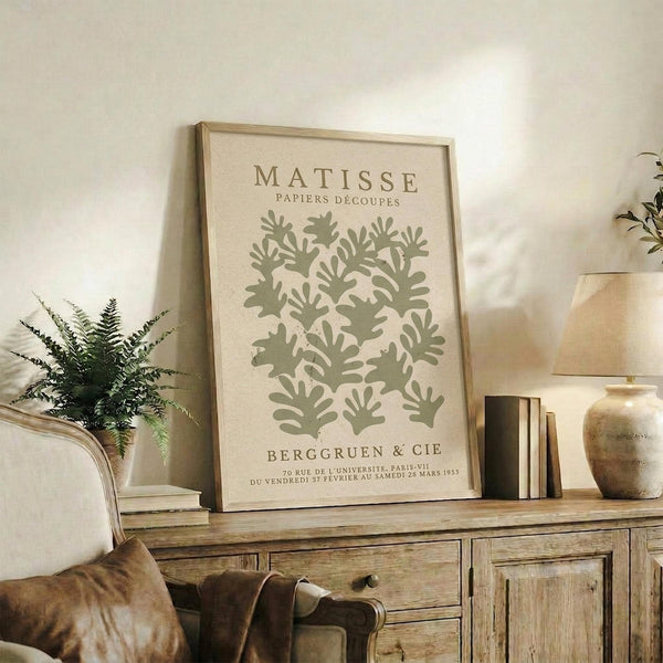 a framed poster of a plant design on a wall, with a lamp and some books on a wooden cabinet below it.