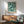 a living room with a large framed poster of a floral pattern hanging on the wall above a wooden cabinet. The poster is titled "William Morris" and is a reproduction of a botanical design. The room has a green armchair and a wooden coffee table, creating a cozy and inviting atmosphere.