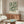 a living room with a large framed poster of a floral pattern on the wall above a wooden cabinet. The poster is titled "William Morris" and "Exhibition Art - Botanical Delight". The room has a white sofa, a glass coffee table, and a lamp on a side table.