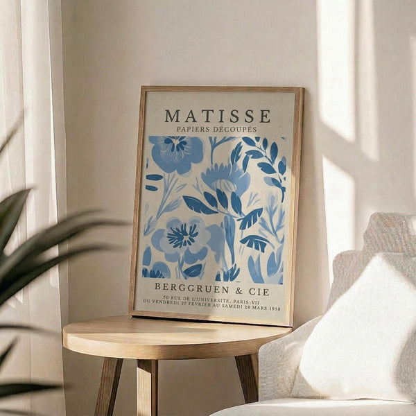 a framed poster of the Métisse art movement, with a floral design in blue and white colors. The poster is displayed on a wooden table, and there is a white chair visible in the background.
