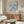 a living room with a large framed poster of the Métisse paper series hanging on the wall above a wooden cabinet. The poster has a blue floral design. The room also includes a white sofa, a glass coffee table, and a lamp on a side table.