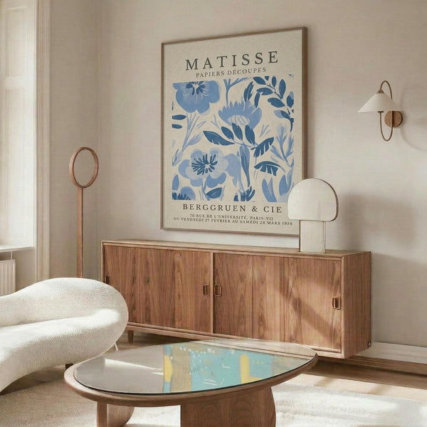 a living room with a large framed poster of the Métisse paper series hanging on the wall above a wooden cabinet. The poster has a blue floral design. The room also includes a white sofa, a glass coffee table, and a lamp on a side table.