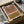 May include: Framed geometric abstract art print featuring terracotta and mustard yellow color blocks with black concentric arch lines, displayed in black frame inside cardboard shipping box with protective tissue paper and wooden dowel tubes visible nearby.