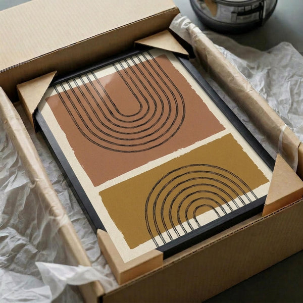 May include: Framed geometric abstract art print featuring terracotta and mustard yellow color blocks with black concentric arch lines, displayed in black frame inside cardboard shipping box with protective tissue paper and wooden dowel tubes visible nearby.