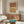 May include: Modern living room featuring a framed abstract print with terracotta and mustard arches on beige wall above wooden credenza. White curved chair, brass floor lamp, marble table lamp, and colorful glass coffee table complete the mid-century styled space with neutral walls and natural lighting.