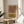 May include: Framed abstract art print featuring geometric rainbow arches in terracotta orange and mustard yellow color blocks with black line details, displayed on a round wooden side table next to a beige upholstered chair. Natural sunlight streams across the neutral-toned room setting with a plant visible in the corner.