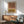 May include: Abstract geometric wall art featuring two arched designs - an upside-down arch in terracotta orange and a rainbow arch in mustard yellow, both with black concentric lines on cream background. Wooden frame hangs above mid-century modern furniture including walnut credenza, sage green upholstered armchair with wooden frame, small round coffee table, and white ceramic table lamp. Warm sunlight streams across white walls.