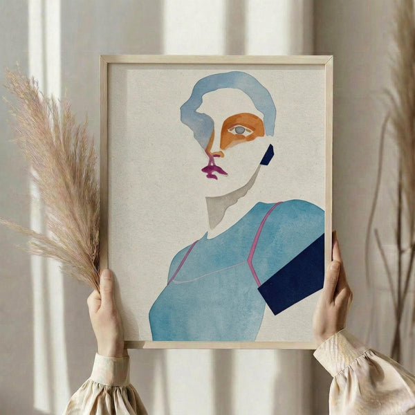 May include: Person holding framed abstract portrait artwork featuring stylized face with blue hair, orange and pink facial features, single visible eye, wearing light blue and dark blue geometric clothing. Dried wheat stems held in other hand. Light wooden frame against neutral cream curtain background.