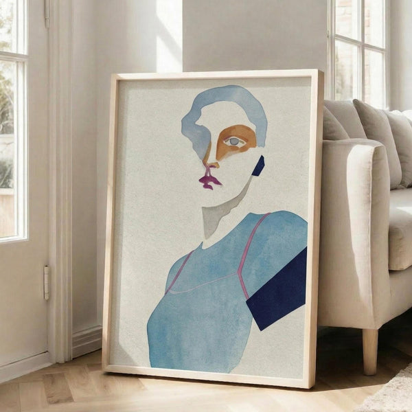 May include: Framed abstract portrait artwork leaning against white wall next to beige upholstered chair. The minimalist illustration features a stylized face with blue hair, orange and pink facial features, single visible eye, and geometric blue and navy clothing shapes. Light wooden frame on hardwood floors in bright, airy room with large windows.