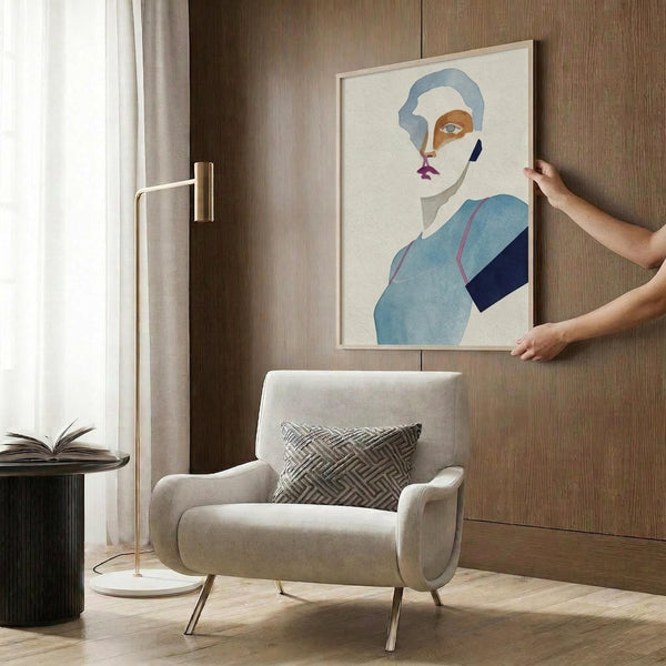 May include: Person's hands positioning a framed abstract portrait print on wood-paneled wall above beige upholstered chair with geometric pillow. Print features stylized face in blue, orange and white tones with modern minimalist design. Room setting shows brass floor lamp, side table with open book, and neutral contemporary decor.
