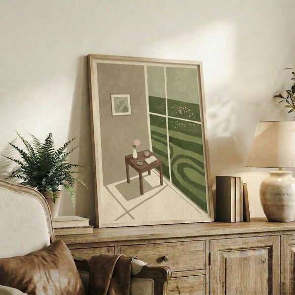 May include: Framed art print displayed on rustic wooden sideboard showing interior room scene with small table, window view of rolling green hills, and wall art. Light wooden frame, muted green and beige tones. Styled with fern plant, ceramic vase, books, and leather sofa visible in cozy living room setting.