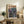 May include: Framed poster displayed on rustic wooden console table in cozy living room setting. Artwork features anime-style illustration of soldier and mech robot in neon-lit cityscape with Japanese text overlay. Scene shows warm lighting, decorative plant, ceramic vase, books, and beige upholstered furniture creating homey atmosphere.