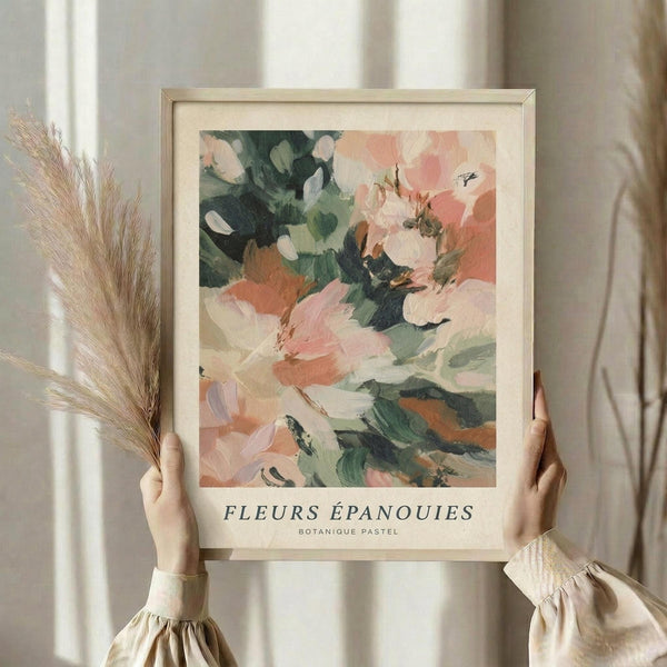 May include: Person in beige ruffle-sleeve top holding framed floral art print and dried wheat stems against white curtained window. The artwork features abstract blooming flowers in soft coral pink, sage green, and cream brushstrokes with "FLEURS ÉPANOUIES BOTANIQUE PASTEL" text below in elegant typography, displayed in light wood frame.