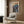 May include: Framed anime-style poster mounted on wood-paneled wall above beige upholstered armchair with geometric throw pillow. Artwork shows figure in military uniform facing large mech robot in futuristic cityscape with neon lighting. Japanese text in orange and teal colors at top. Modern reading chair with gold legs, brass floor lamp, and dark round side table with open book complete the contemporary room setting.