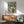 May include: Framed poster showing armored figure with umbrella walking through rainy futuristic city street with giant mecha silhouette looming overhead, Japanese text at top, displayed above wooden sideboard with white ceramic lamp in modern living room with mint green upholstered chair and natural lighting casting shadows on white wall.