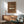 May include: Brown vintage-style poster featuring classic white Porsche 911 sports car with "PORSCHE 911 CLASSIC" text and "1964" date, displayed in wooden frame above mid-century modern walnut credenza with white ceramic table lamp, next to sage green upholstered armchair with wooden arms in bright sunlit room with white walls and dramatic window shadows.