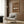 May include: Framed brown poster showing white vintage sports car with "911 CLASSIC" and "1964" text, displayed on wood paneled wall above beige upholstered armchair with geometric patterned pillow, brass floor lamp, and dark round side table with open book in modern living room setting