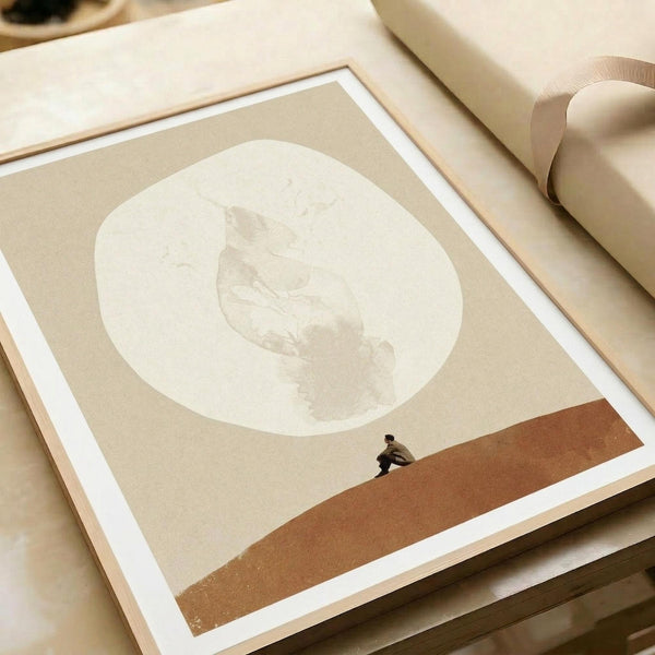 A minimalist poster print featuring a small silhouetted figure walking along a rust-colored ground beneath a large pale circular moon against a cream background. The artwork is displayed in a simple wooden frame on textured paper, photographed at an angle with neutral beige wrapping paper and ribbon visible in the scene.