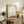 May include: Framed surrealist artwork displaying a large pale moon above a rust-colored desert landscape with a small silhouetted figure, shown in a neutral-toned living room setting on a rustic wooden sideboard alongside books, a ceramic vase, and fern plant, with warm natural lighting and beige upholstered furniture visible in the scene.