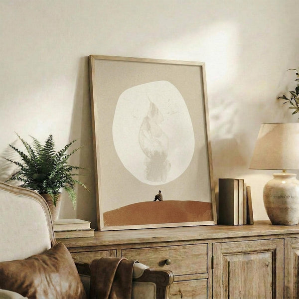 May include: Framed surrealist artwork displaying a large pale moon above a rust-colored desert landscape with a small silhouetted figure, shown in a neutral-toned living room setting on a rustic wooden sideboard alongside books, a ceramic vase, and fern plant, with warm natural lighting and beige upholstered furniture visible in the scene.