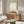 May include: Framed beige and white abstract poster with oval shape and subtle figure silhouette, displayed above walnut wood sideboard in modern living room. White mushroom table lamp, curved cream chair, oval glass coffee table with colorful abstract surface, and tall brass mirror on stand complete the neutral-toned Scandinavian-style interior scene.