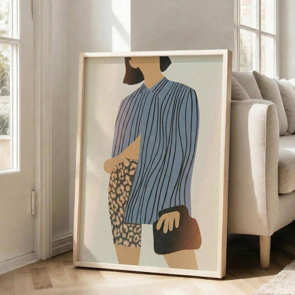May include: Large framed abstract fashion print leaning against white wall in bright living room. Artwork shows minimalist figure in blue striped top and leopard print skirt holding dark purse. Light wood frame, beige sofa visible, natural sunlight streaming through windows onto hardwood floors.