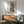 May include: Modern living room with mid-century furniture featuring a framed abstract fashion illustration on white wall. Artwork shows stylized figure in blue striped top and leopard print skirt holding brown handbag. Room includes sage green upholstered chair with wooden arms, walnut credenza with white ceramic lamp, and round coffee table. Natural sunlight creates diagonal shadows across the scene.