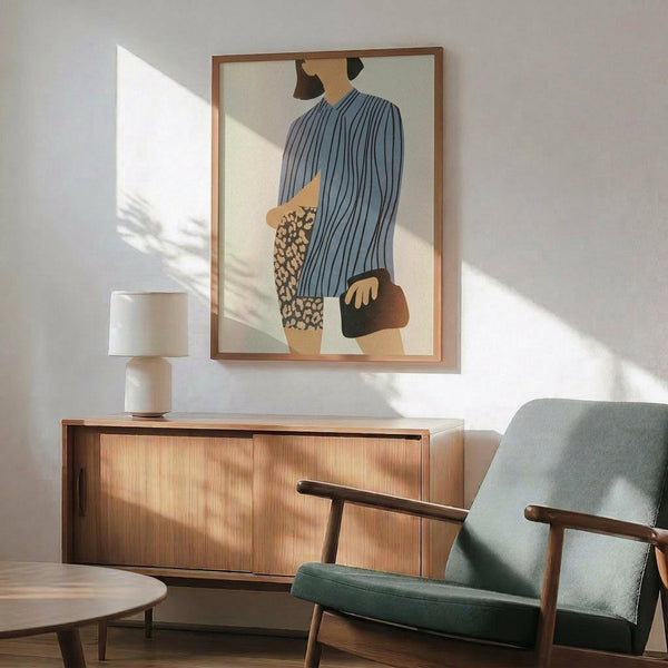 May include: Modern living room with mid-century furniture featuring a framed abstract fashion illustration on white wall. Artwork shows stylized figure in blue striped top and leopard print skirt holding brown handbag. Room includes sage green upholstered chair with wooden arms, walnut credenza with white ceramic lamp, and round coffee table. Natural sunlight creates diagonal shadows across the scene.