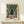 May include: Framed poster displaying retro-style anime artwork featuring a silhouetted figure in a trench coat walking down a rainy city street with a large green mecha robot looming overhead. Japanese text appears at the bottom in orange lettering. The poster has a vintage comic book aesthetic with teal, orange, and green colors. Displayed on a light wood shelf with a small wrapped gift beside it against a white wall.