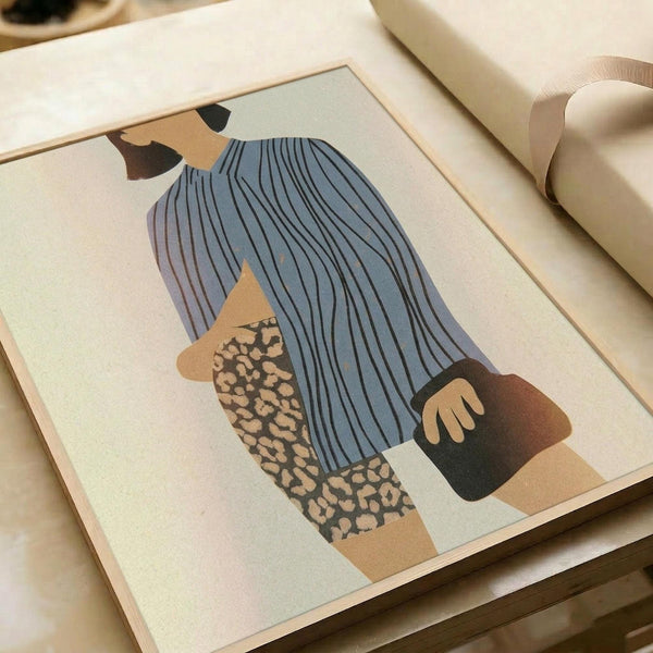 Stylized illustration of a seated figure with long blue hair wearing a striped blue top and leopard print pants, rendered in muted tones of blue, beige, and brown. The minimalist artwork is displayed in a light wooden frame on a cream-colored surface next to rolled paper, showing warm natural lighting and soft shadows.
