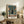 May include: Framed vintage-style Japanese movie poster featuring a silhouetted figure walking through a rainy cyberpunk city street with a large mecha robot looming overhead. The artwork uses muted green and orange tones with detailed urban architecture. The poster sits on a rustic wooden sideboard next to a potted fern, ceramic vase, and books in a cozy living room setting with a beige armchair visible.