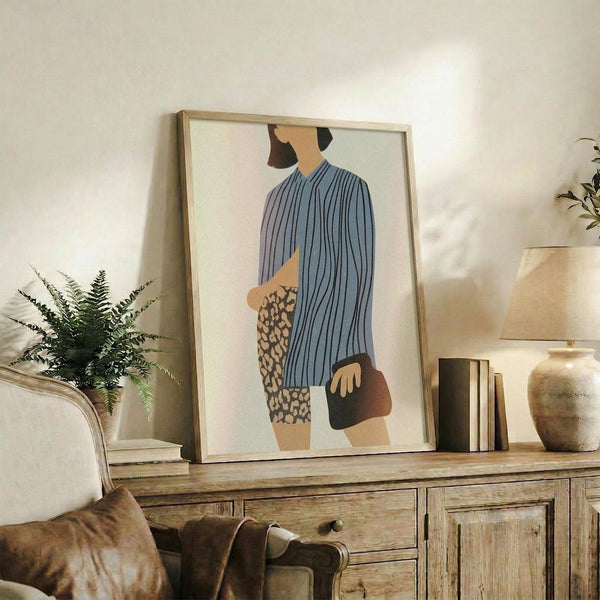 May include: Framed abstract figure print displayed on rustic wooden sideboard in neutral living room setting. Artwork shows stylized person in blue striped top and leopard print skirt against cream background. Scene includes fern plant, ceramic vase, books, and beige upholstered furniture with warm natural lighting.