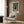 May include: Framed vintage-style poster featuring a silhouetted figure standing before a towering mech robot in a rainy cyberpunk cityscape with neon signs and Japanese text, displayed on wood-paneled wall above cream upholstered armchair with geometric patterned pillow, beside brass floor lamp and small round table with open book