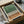 Abstract art print with vertical black lines creating a forest silhouette effect against green and tan gradient background. The minimalist poster is displayed in a wooden frame inside an open cardboard shipping box with protective tissue paper. Two wooden tubes and a dark container are visible nearby on white fabric, showing the careful packaging for shipping.