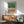 May include: Abstract watercolor artwork with green wash at top flowing into black vertical lines resembling grass or reeds, with warm brown wash at bottom, displayed in light wood frame above mid-century modern walnut credenza with ceramic table lamp, next to sage green upholstered armchair with wooden arms in sunlit room with white walls