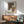 May include: Abstract geometric poster with brown, beige, and dotted pattern shapes displayed in wooden frame above mid-century modern credenza. Scene shows mint green upholstered chair with wooden arms, white ceramic table lamp, and natural sunlight casting shadows on white wall in contemporary living room setting.