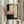 May include: Person in cream ruffle-sleeve top holding framed abstract artwork featuring geometric shapes in pink, cream, black and gray tones with vertical row of black dots down center, dried wheat stems visible to left, soft natural lighting from window in background