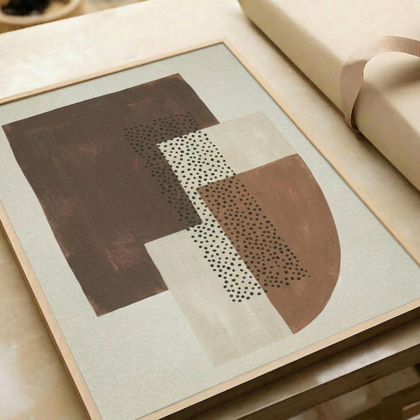 May include: Framed abstract art print with earthy geometric shapes in brown and terracotta tones, featuring dotted stippled textures overlaying curved and angular forms, displayed on a light wooden surface next to neutral colored paper rolls in soft natural lighting