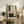 May include: Abstract art print in light wood frame displayed on rustic wooden sideboard in modern living room. Artwork features geometric blocks in pink, beige, black and gray tones with vertical dotted line detail. Scene includes fern plant, stacked books, ceramic vase, and cream upholstered chair with brown leather throw blanket.